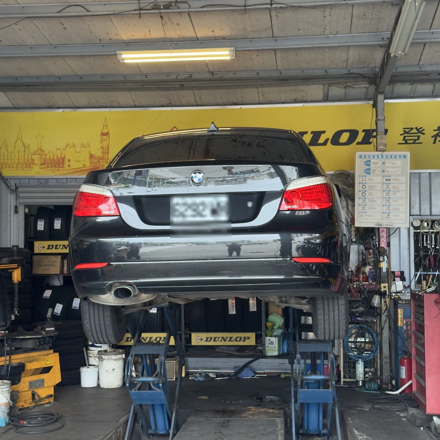 黑色 BMW E60 520d 被架在輪胎行的舉升機上，從車尾方向拍攝