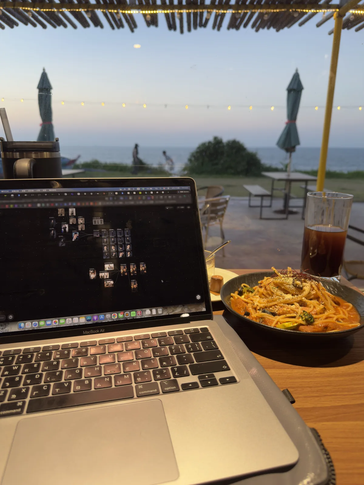 傍晚的戶外座位亮起串燈，桌上是 MacBook、義大利麵與冰美式