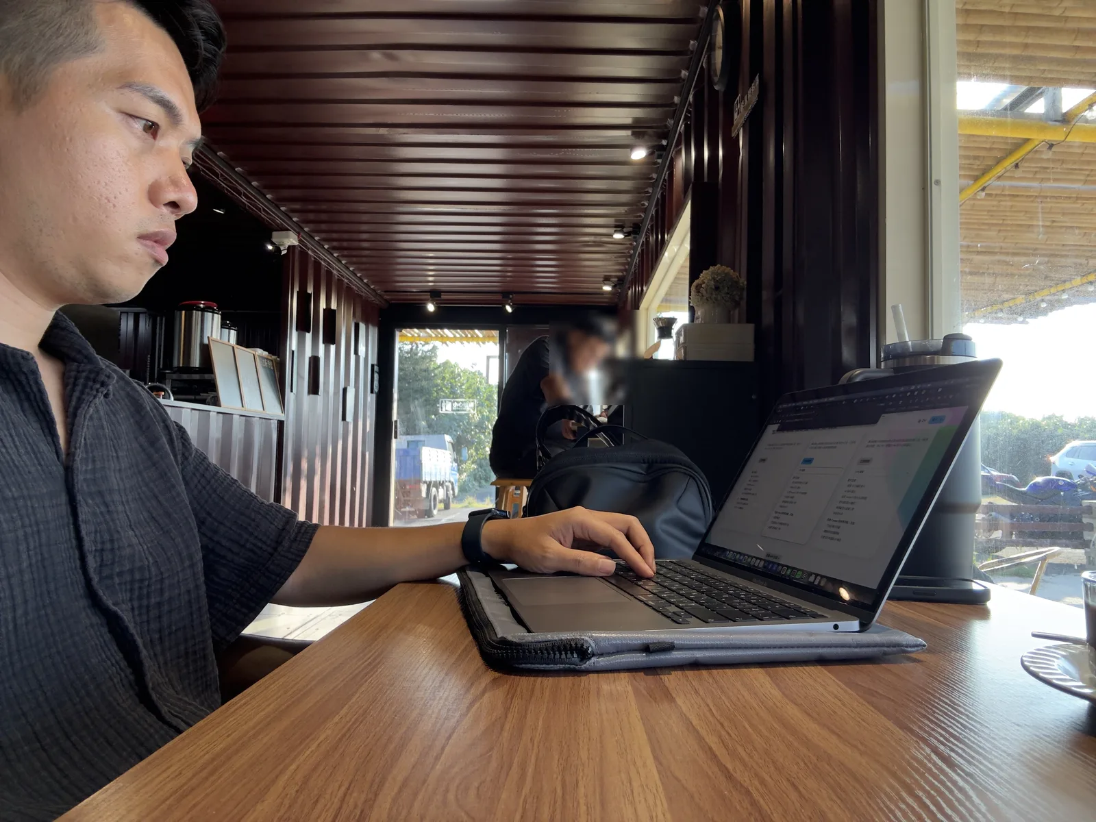 Jimmy 坐在貨櫃屋室內的長桌前使用 MacBook 工作，室內是深色貨櫃內裝